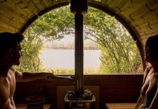 Pareja relajándose en una sauna acogedora con vista al lago desde un alojamiento de glamping en la naturaleza.