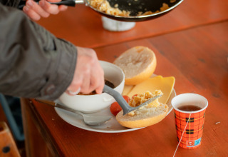 Una persona prepara desayuno con pan, queso, huevos revueltos y té en una mesa de alojamiento glamping.