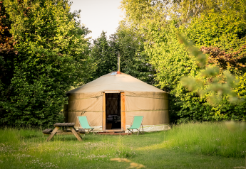 En hyggelig yurt ved RØSTIG Dorst i Holland, omgivet af grønne træer med stole og et picnicbord foran.