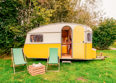 Gelber Vintage-Wohnwagen mit offener Tür, zwei Stühlen und Kiste im Grünen bei RØSTIG Dorst, Niederlande.