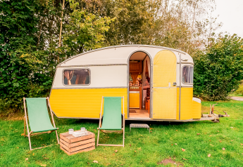 Caravana retro amarilla con puerta abierta, dos sillas y mesa en césped en RØSTIG Dorst, Países Bajos.