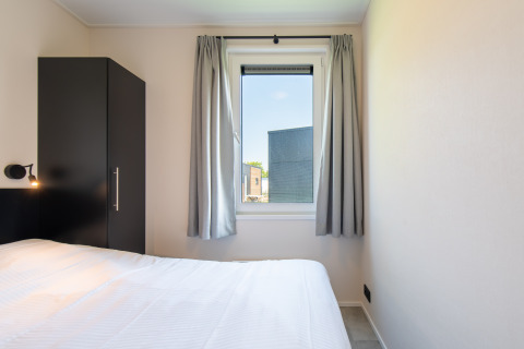 Dormitorio en Wood Lodge con cama individual, armario negro y ventana con cortinas grises en Kompas Camping Nieuwpoort.