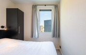 Slaapkamer in Wood Lodge met eenpersoonsbed, zwarte kast en raam met grijze gordijnen op Kompas Camping Nieuwpoort.
