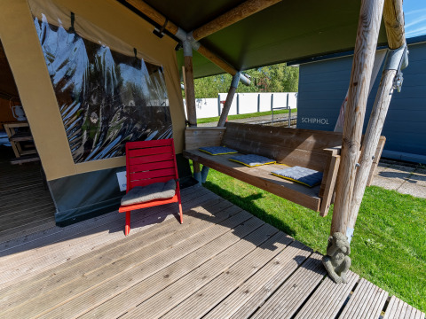 Coin salon extérieur à la Safari tent Heeg à Marina Heeg, Pays-Bas, avec banc en bois et chaise rouge.