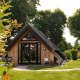 Houten tiny house genaamd Wellness Lodge met terras, grasveld en veel bomen op de achtergrond.