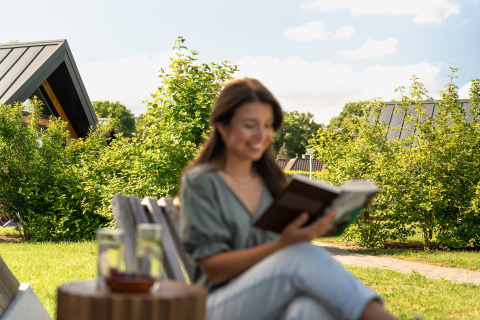 Woman reading a book outdoors at the Wellness Lodge tiny house in Holiday Park De IJsvogel, Netherlands.