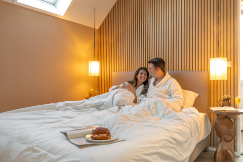 Coppia in accappatoi fa colazione a letto nella Wellness Lodge al Holiday Park De IJsvogel, Paesi Bassi.