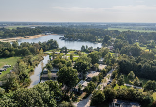 Vue aérienne d'un hébergement glamping au bord d’un lac, entouré de verdure et d’arbres par temps ensoleillé.