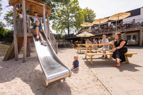 Niños jugando en un tobogán y adultos sentados en mesas con sombrillas en alojamiento glamping.