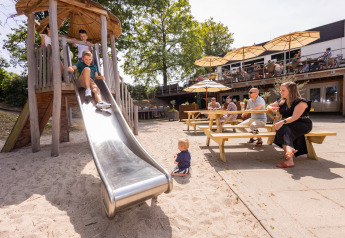 Kinder rutschen auf einer Rutsche, Erwachsene sitzen an Tischen mit Sonnenschirmen in einer Glamping-Anlage.