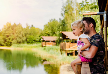 Vater und Kind blicken von einer Glamping-Unterkunft aus auf einen See, umgeben von Bäumen und Hütten.