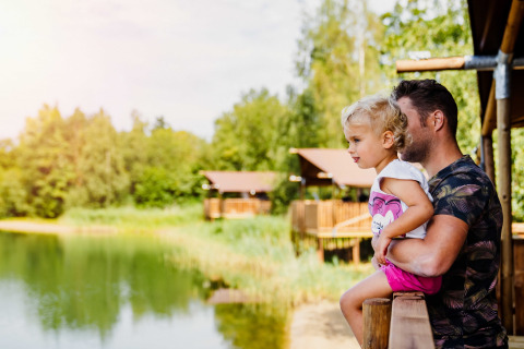 Padre sostiene a su hijo pequeño en una terraza, contemplando un lago en un glamping rodeado de cabañas y naturaleza.