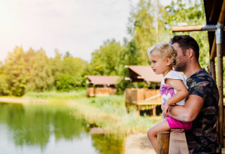 Padre sostiene a su hijo pequeño en una terraza, contemplando un lago en un glamping rodeado de cabañas y naturaleza.
