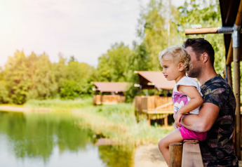 Un père tient son enfant sur une terrasse, admirant un lac près des cabanes, dans un site de glamping en pleine nature.
