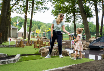 Mor og datter spiller minigolf sammen i et udendørs glampingområde omgivet af grønne træer.