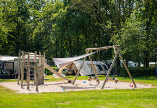 Parco giochi con altalene e scivolo in un glamping, circondato da tende e vegetazione lussureggiante.