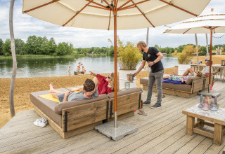 Huéspedes descansan bajo sombrillas en una terraza junto al lago, disfrutando de unas vacaciones glamping.