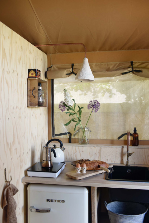 Petite cuisine rétro sous tente safari avec pain, œufs, bouilloire, vase de fleurs et réfrigérateur.