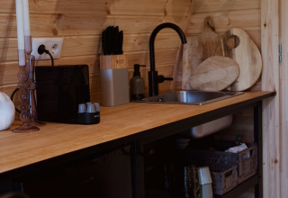 Piano cucina rivestito in legno con rubinetto nero, cestini appesi e ripiani in un glamping pod nei Paesi Bassi.