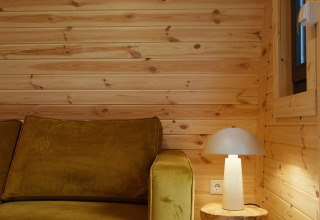 Knus hoekje met mosterdgele zetel, houten tafeltje en lamp in Glamping Pod met Sauna, Nederland.