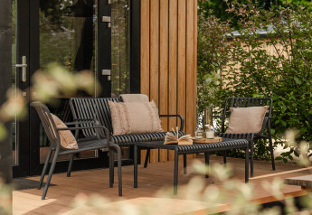 Terraza exterior de una tiny house con muebles de jardín negros modernos, cojines y libros abiertos en la mesa.