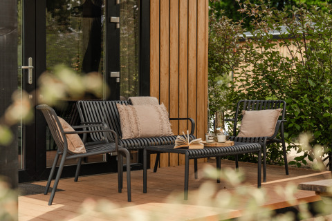 Patio esterno di una tiny house con mobili da giardino neri moderni, cuscini e libri aperti sul tavolino.