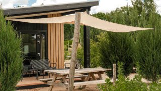 Outdoor picnic table and shade sail in front of Sand Lodge Sauna tiny house at Holiday Park De IJsvogel, Netherlands.