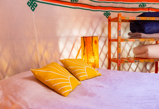 Interior de una yurta con una cama de almohadas amarillas, estantería colorida y luz cálida tradicional.