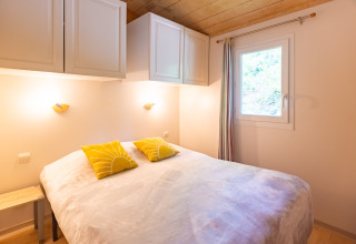 Dormitorio en chalet de Camping Des Randonneurs, Francia, con cama doble, ventana y cojines amarillos.
