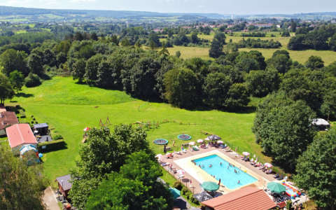 Aerial view of a glamping site featuring a pool, green fields, playground, and surrounding forest scenery.
