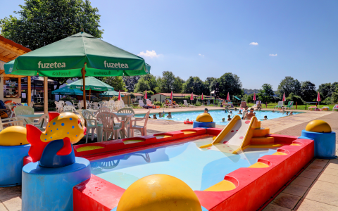Zona de piscina en glamping con área infantil, tobogán pequeño y mesas bajo sombrillas Fuzetea verdes.