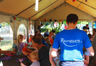 Niños y adultos haciendo manualidades juntos bajo una carpa en un glamping, guiados por un animador.