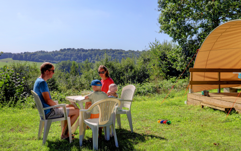 Family enjoys outdoor glamping at a table with children, plastic chairs, and scenic countryside views.