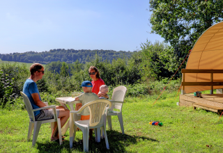 Gezin geniet van glamping buiten aan een tafel met kinderen, plastic stoelen en fraai uitzicht op natuur.