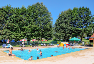 Outdoor swimming pool at a glamping accommodation with guests, sunbeds, parasols, and leafy trees nearby.