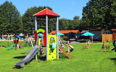 Kinder spielen auf einem Spielplatz mit Rutsche in einer Glamping-Unterkunft mit grüner Wiese und Sonnenschirmen.