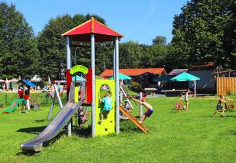 Kinderen spelen op een speeltuin met glijbaan bij een glampingverblijf met groene gazons en parasols.