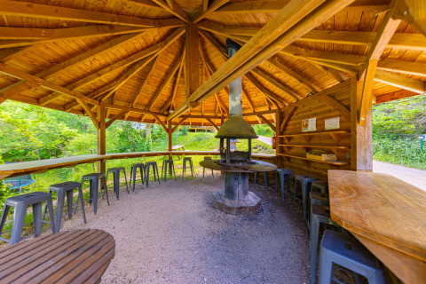 Pabellón cubierto de madera con barbacoa central y taburetes en Camping Des Randonneurs, Occitania, Francia.