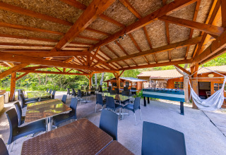 Overdekte buiteneethoek bij Camping Des Randonneurs, Occitanië, Frankrijk, met tafels, stoelen, hangmat en voetbaltafel.
