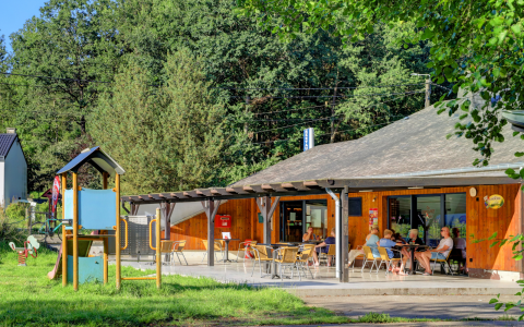 Terrasse d'un café en hébergement glamping, avec invités, aire de jeux et forêt en arrière-plan.