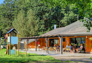 Buitenzitje bij een glampingverblijf met gasten, kinderspeeltuin en bomen op de achtergrond.