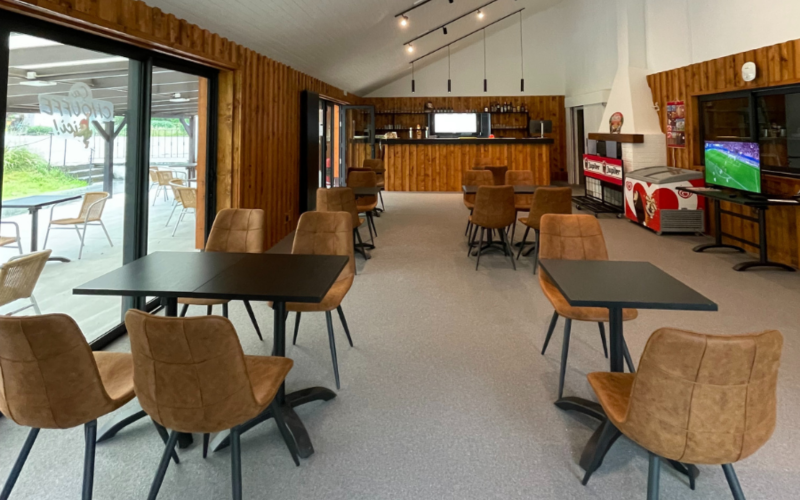 Espace lounge moderne dans un hébergement glamping avec chaises marron, tables noires et grande baie vitrée.