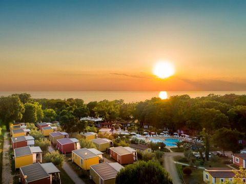 Zonsondergang boven chalets en bomen bij zee, met een zwembad en de zee op de achtergrond.