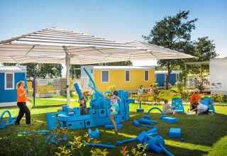 Bambini giocano con grandi blocchi blu in gommapiuma vicino a cabine colorate sotto una tettoia.