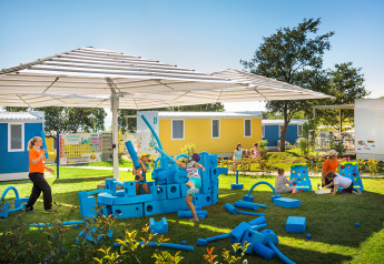 Bambini giocano con grandi blocchi blu in gommapiuma vicino a cabine colorate sotto una tettoia.