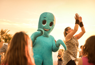 I bambini ballano all'aperto al tramonto con una persona in costume da polpo blu durante una festa allegra.