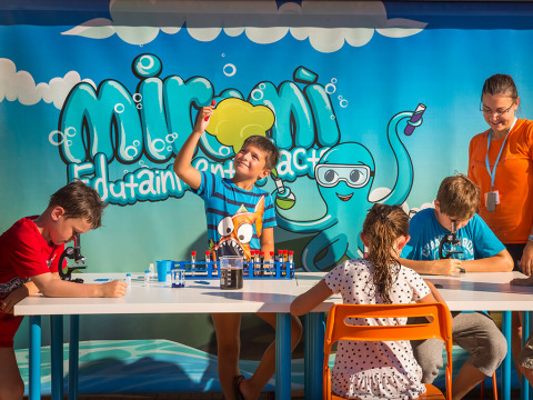 Kinderen voeren wetenschappelijke proefjes uit aan een tafel met een begeleider voor een kleurrijke cabine.