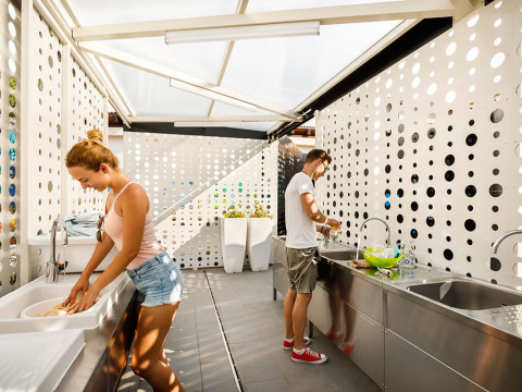 Dos personas lavan platos en una moderna zona de cocina con paredes blancas perforadas de un parque vacacional.
