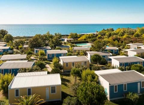 Des cabanes colorées entourées de verdure sont installées près de la mer sous un ciel dégagé.