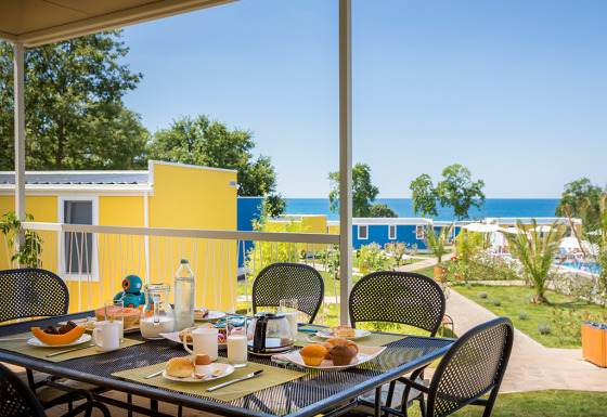 Ontbijttafel buiten met eten en drinken en zicht op kleurrijke chalets en zee in Miramí Family Village.